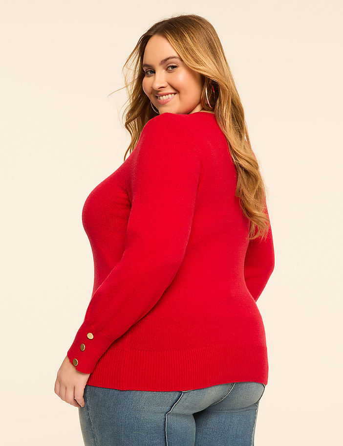 A woman in a red sweater and jeans smiles over her shoulder against a plain, light background.