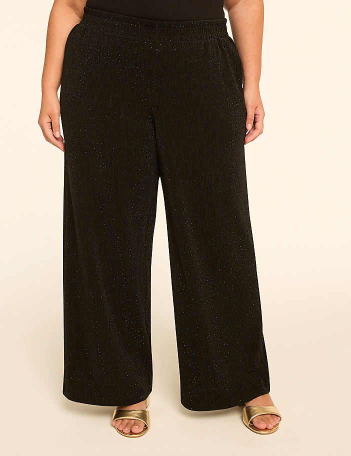 A person stands in wide-leg black sparkly pants and gold sandals against a plain cream background.