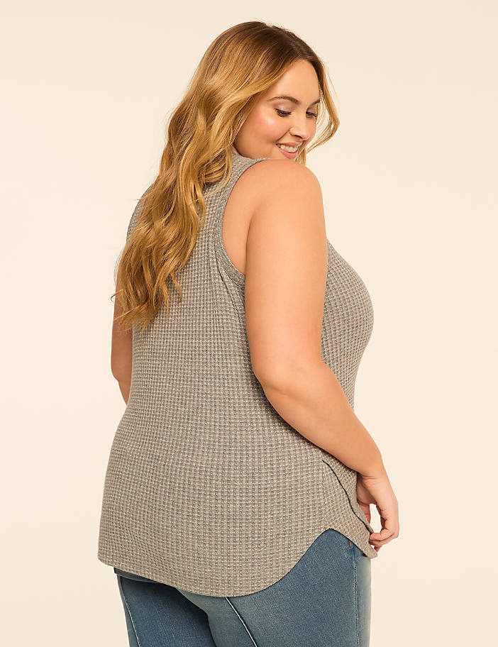 A woman in a gray waffle-knit tank top and blue jeans smiles over her shoulder against a plain, light-colored background.