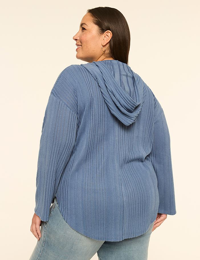 A woman in a blue ribbed hoodie and jeans stands against a plain, light-colored background.