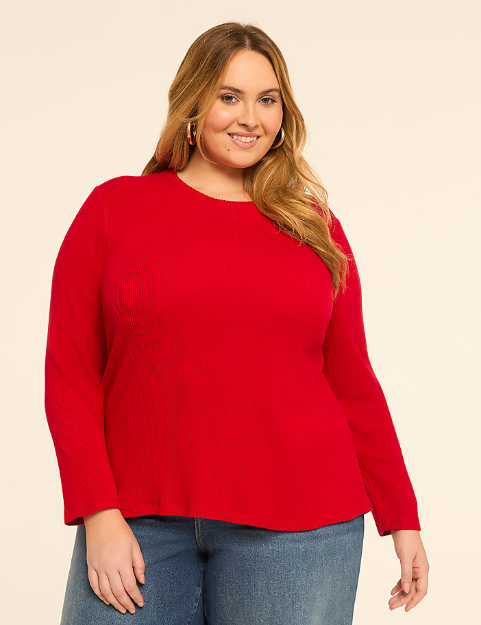A smiling woman in a red sweater and blue jeans stands against a plain, light-colored background.