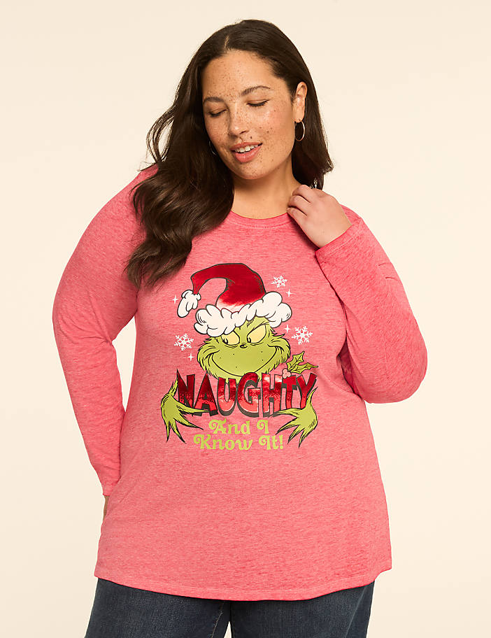 A woman in a pink long-sleeved shirt with a Grinch graphic stands against a plain, light-colored background.