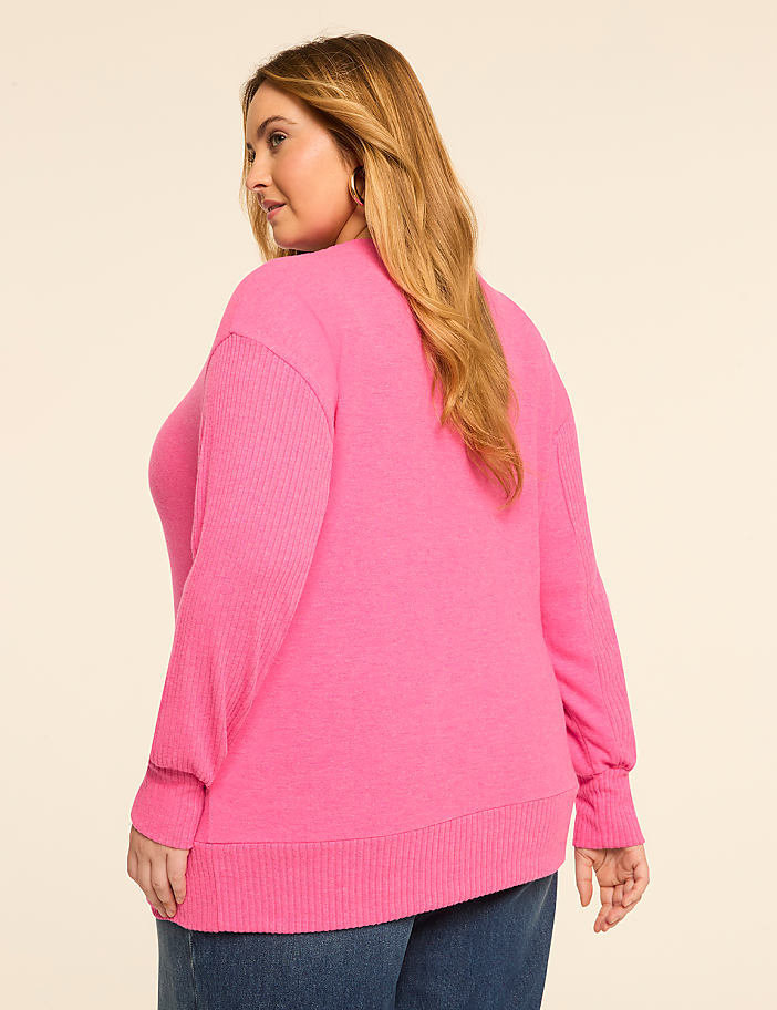 A woman in a bright pink sweater and jeans stands against a plain, light-colored background.