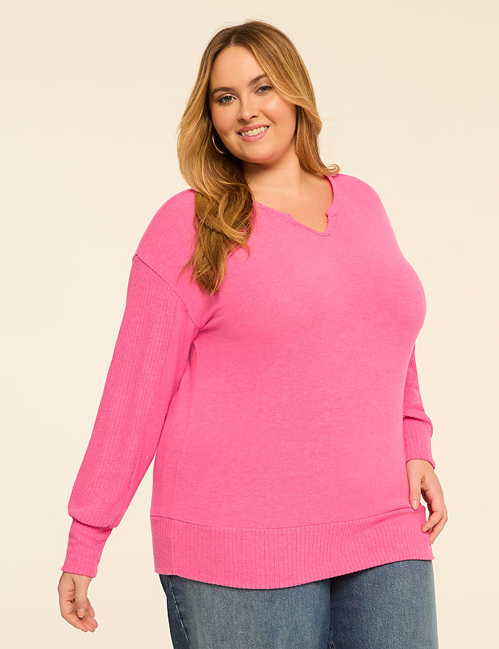 A smiling plus-size woman in a bright pink v-neck sweater and blue jeans stands against a plain, light-colored background.