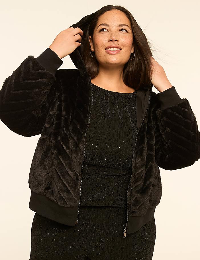 A smiling woman in a black faux fur jacket and sparkly top poses against a plain, light-colored background.