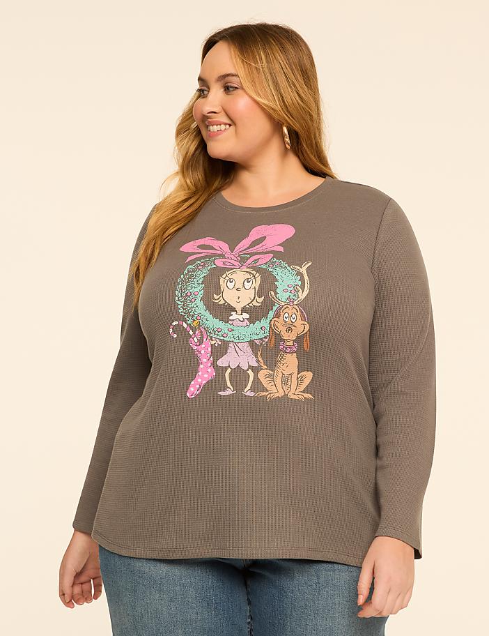 A woman in a grey long-sleeved shirt with a Christmas-themed graphic of Cindy Lou Who and Max the dog stands against a plain beige background.
