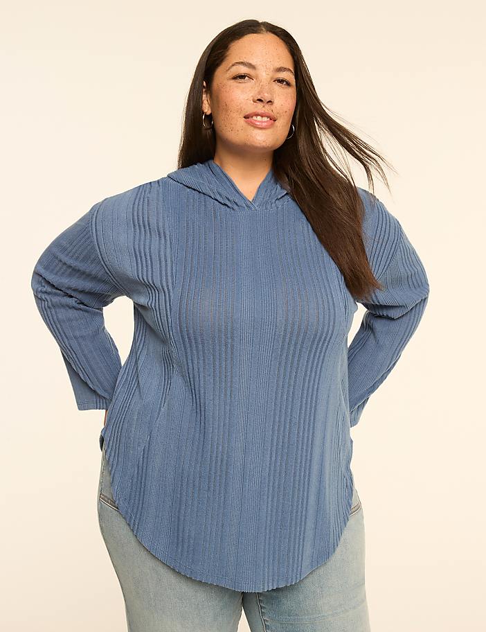 A woman in a blue ribbed hoodie and jeans stands against a plain, light-colored background.
