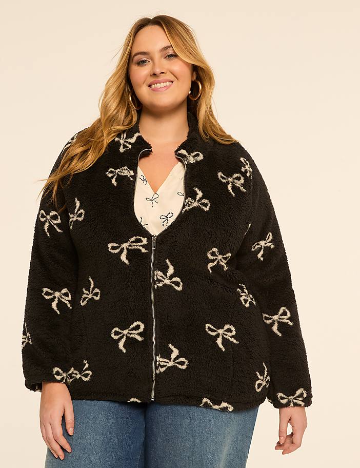 A smiling woman in a fuzzy black jacket with a bow pattern stands against a plain, light-colored background.