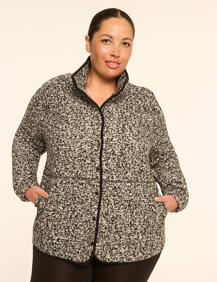 A woman in a speckled jacket with her hands in her pockets stands against a plain, light-colored background.