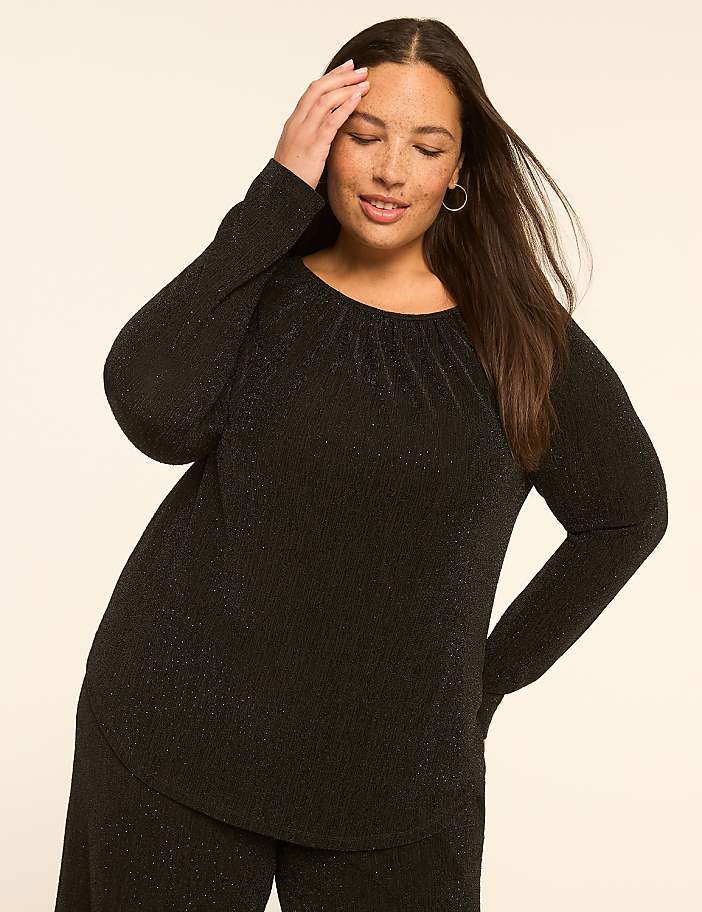 A woman in a black sparkly top and pants poses with her hand on her head against a plain beige background.