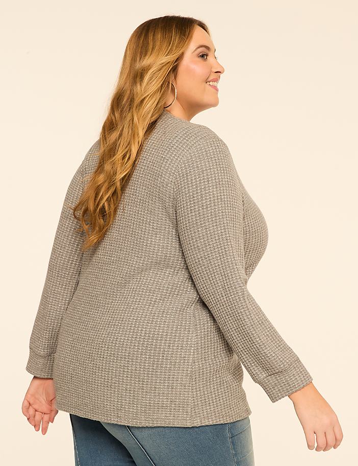 A woman in a gray waffle-knit top and blue jeans stands against a plain, light-colored background.