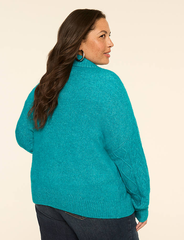 A woman in a teal sweater and jeans stands against a plain, light-colored background.