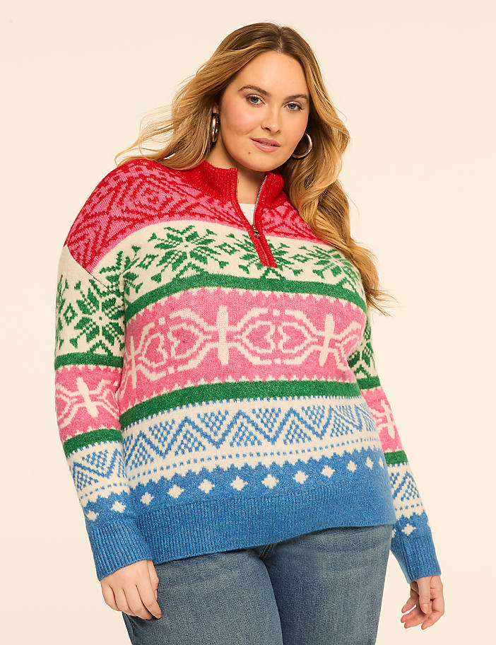 A woman in a colorful, patterned sweater stands against a plain, light-colored background.