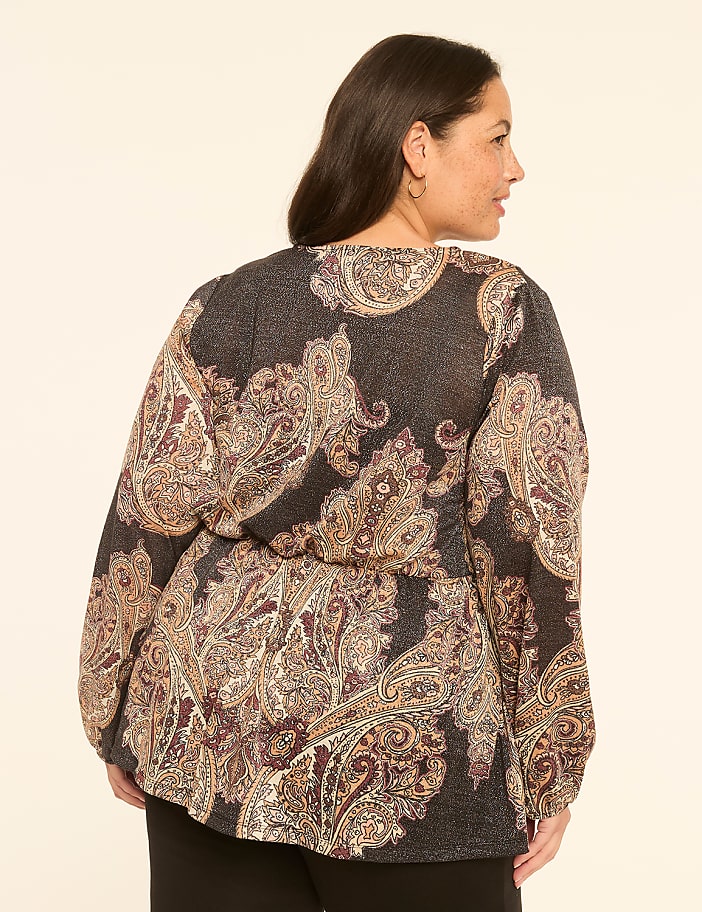 A woman in a paisley-patterned blouse stands against a plain, light-colored background.