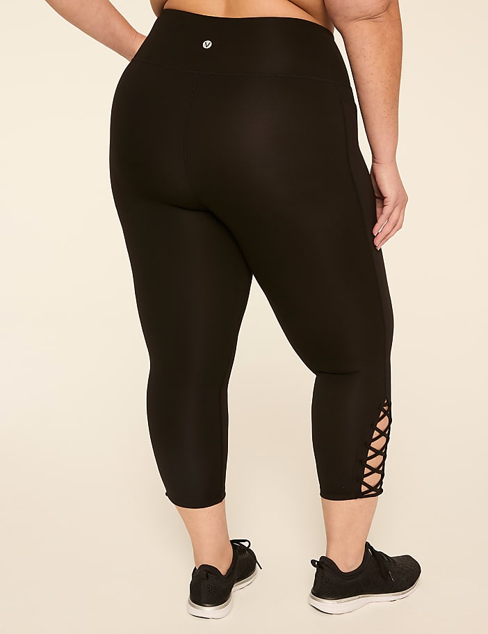 A person wearing black capri leggings with a lattice detail at the ankle stands against a plain cream background.
