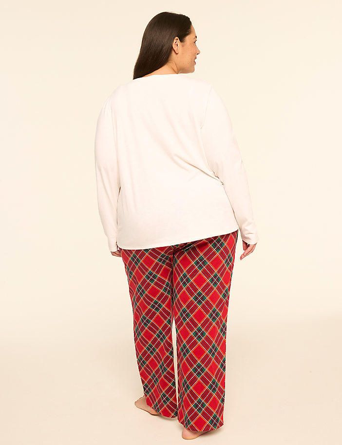 A woman in a white long-sleeved shirt and red plaid pajama pants stands barefoot against a plain, light-colored background.