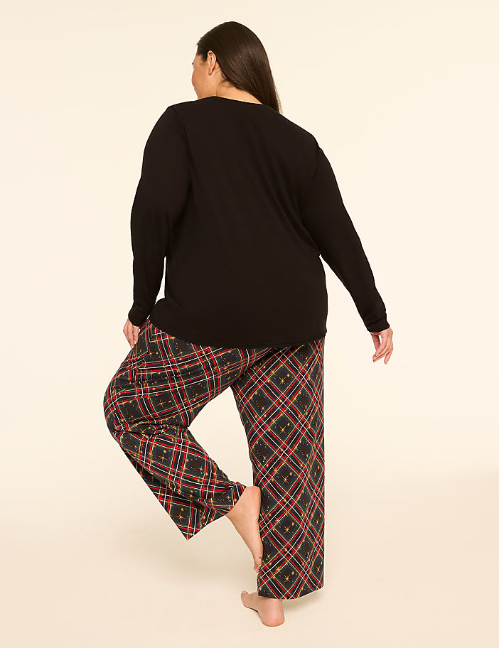 A woman in a black long-sleeved shirt and plaid pajama pants stands on one leg against a plain, light-colored background.