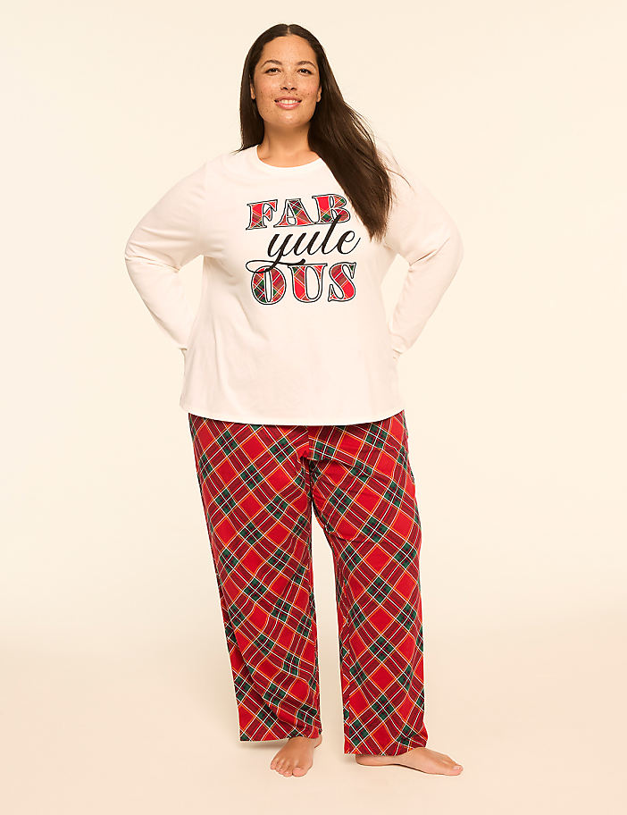 A woman in a white long-sleeved shirt with "Fabulously" printed on it and red plaid pajama pants stands against a plain, light-colored background.