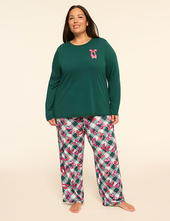 A woman stands in a green long-sleeved shirt with a pink bow graphic and patterned pajama pants against a plain, light-colored background.