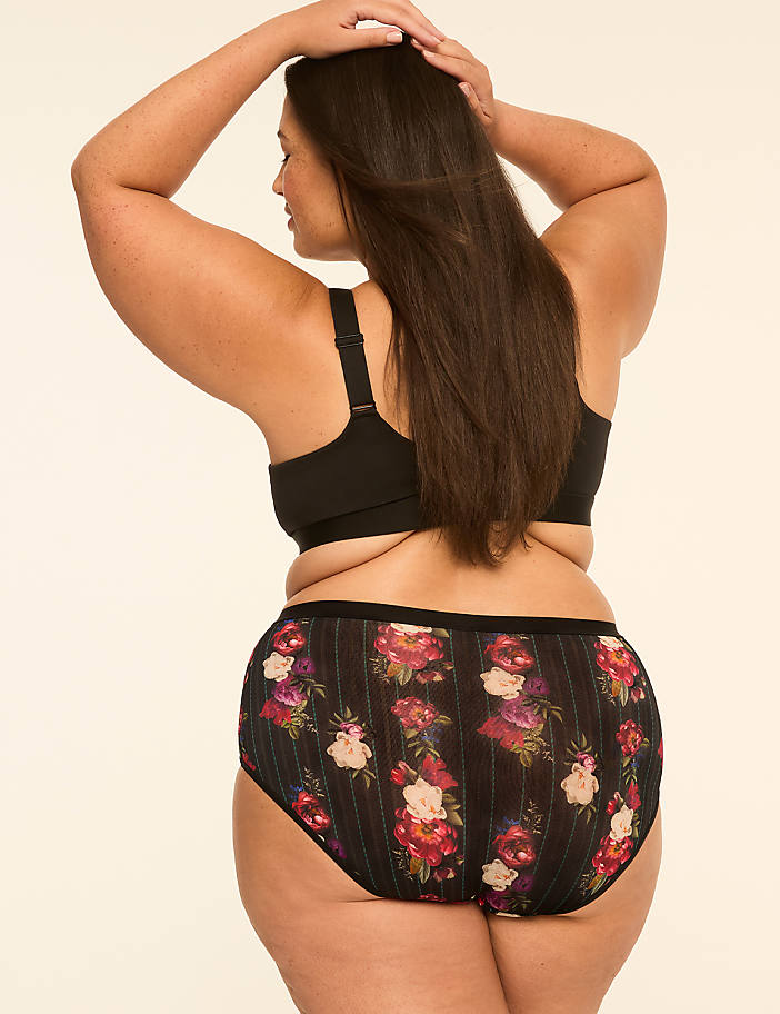 A woman in floral underwear and a black bra stands against a plain, light-colored background with her hands behind her head.