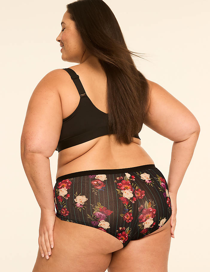 A woman in a black bra and floral patterned underwear stands against a plain, light-colored background.