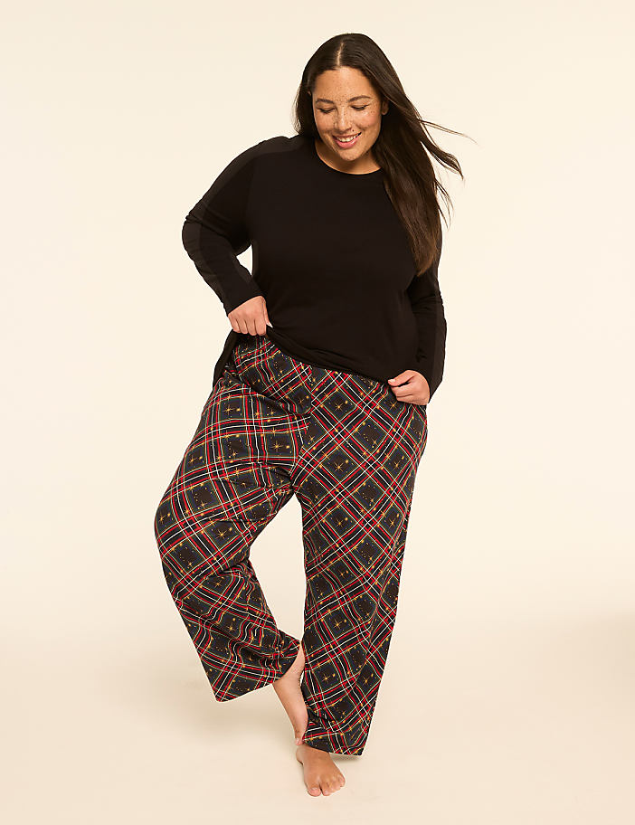 A smiling woman in a black top and plaid pajama pants stands on a plain, light-colored background.