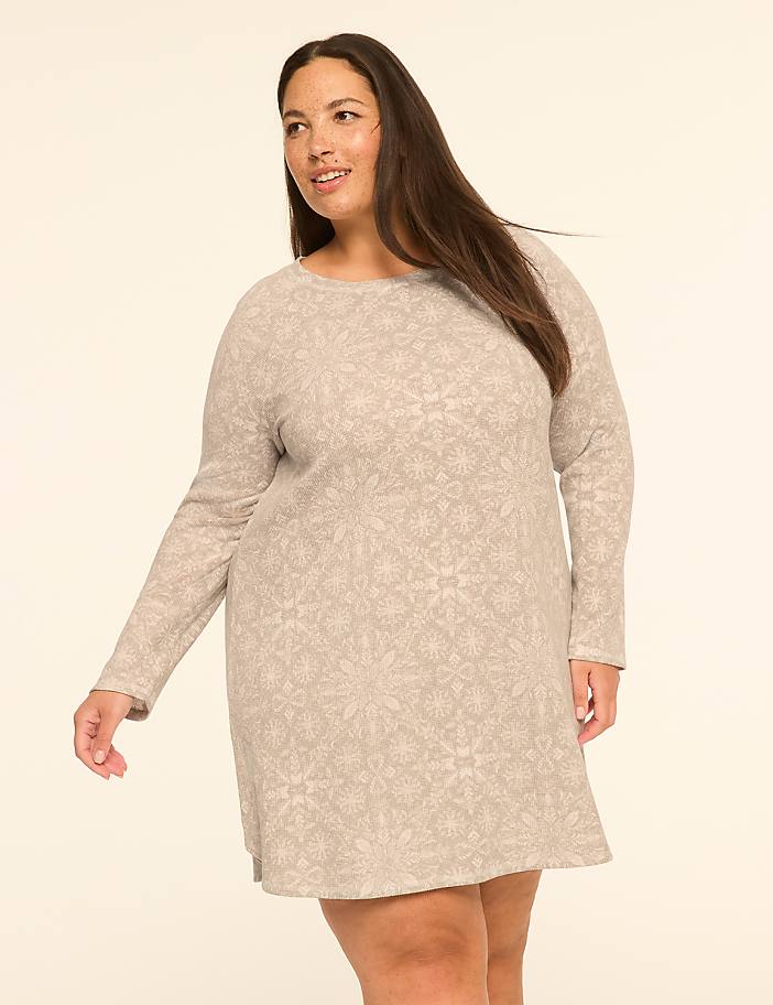 A woman in a patterned dress stands against a plain, light-colored background.