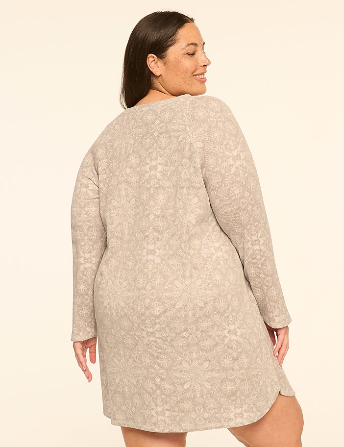 A woman in a patterned dress stands against a plain, light-colored background.
