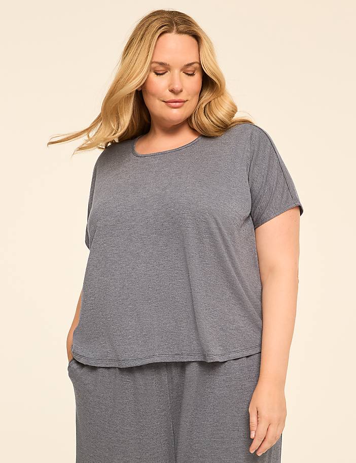 A blonde woman in a grey t-shirt and pants stands against a plain, light-colored background.