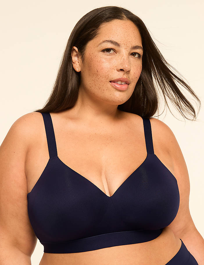 A woman with dark hair and freckles poses in a navy blue bra against a plain, light-colored background.