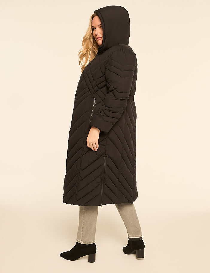 A woman in a black puffer coat and jeans stands against a plain, light-colored background.
