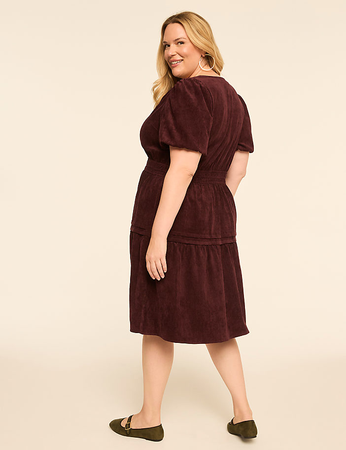 A woman in a burgundy corduroy dress stands against a plain cream background.