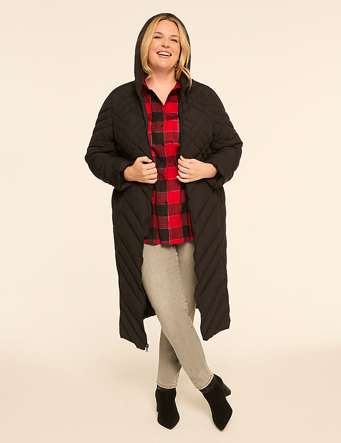 A smiling woman in a black quilted coat and red plaid shirt stands against a plain, light-colored background.