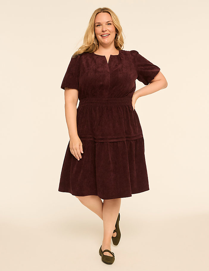 A smiling woman in a burgundy corduroy dress stands against a plain, light-colored background.
