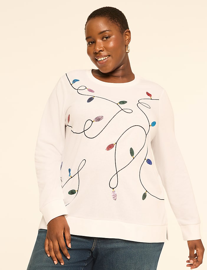 A woman in a white sweatshirt with a string of colorful Christmas lights design smiles against a plain, light-colored background.