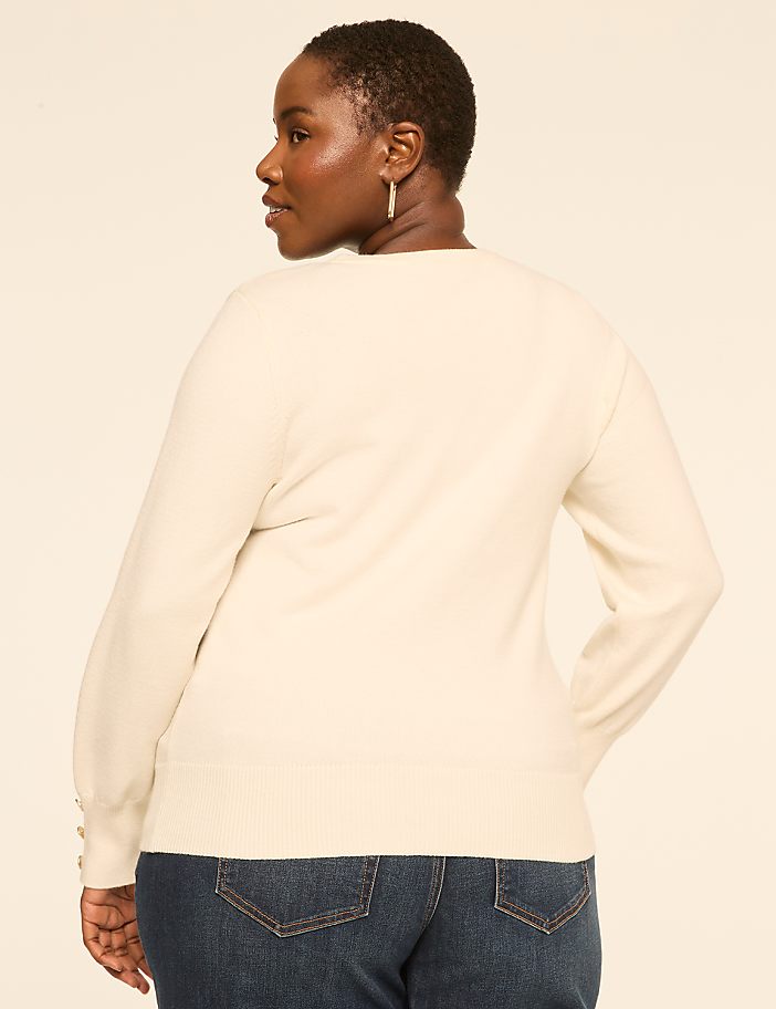A woman in a cream sweater and blue jeans stands against a plain, light-colored background.