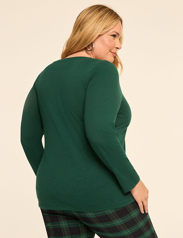 A woman in a green long-sleeved shirt and plaid pants stands against a plain, light-colored background.