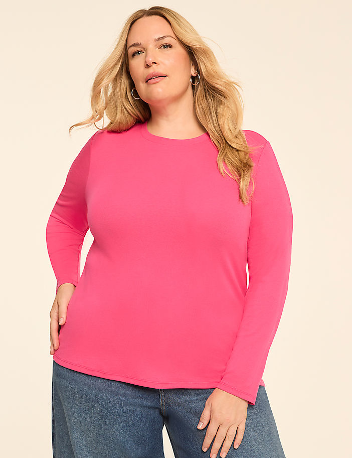 A blonde woman in a pink long-sleeved shirt and blue jeans stands against a plain, light-colored background.