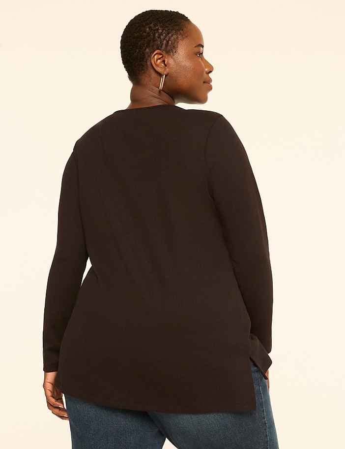 A woman in a black top and blue jeans stands against a plain, light-colored background.