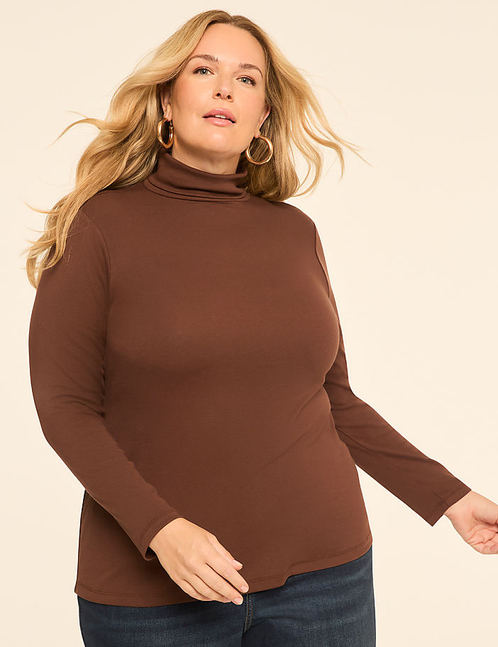 A woman in a brown turtleneck and jeans poses against a plain, light-colored background.