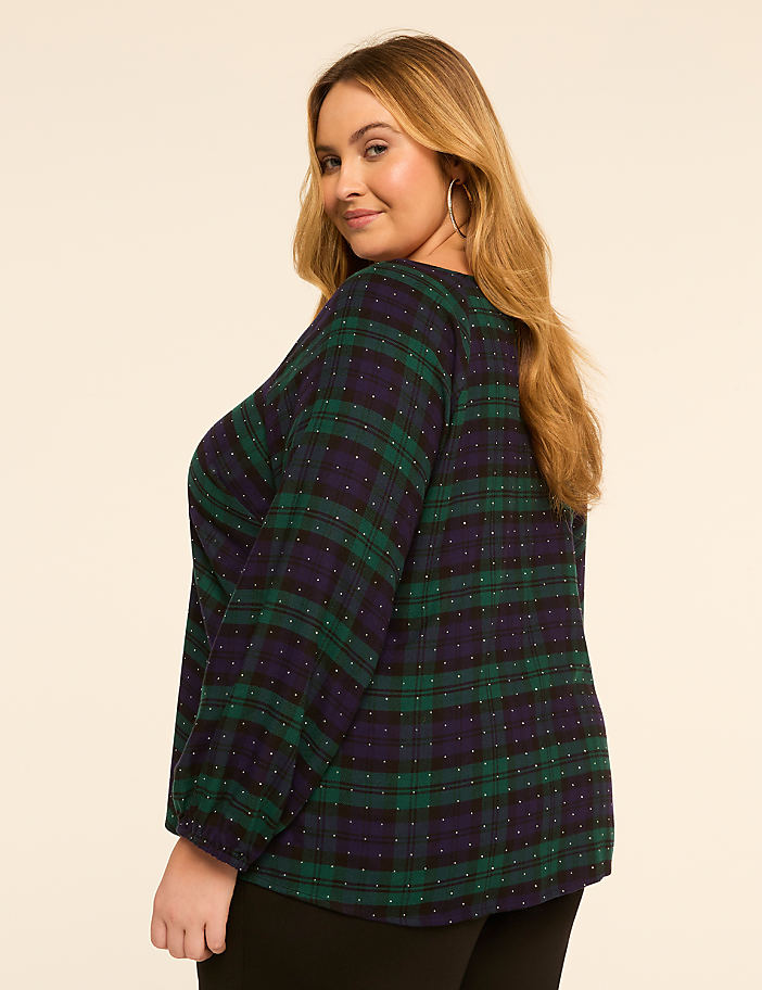 A woman in a green and navy plaid top with white polka dots poses against a plain, light brown background.