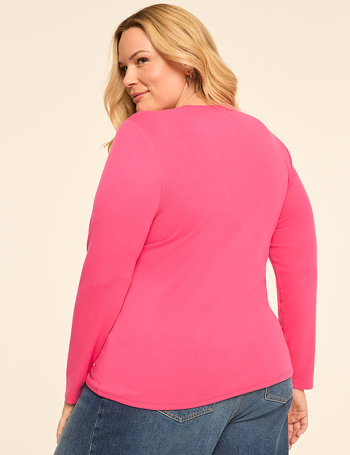A woman in a bright pink long-sleeved shirt and blue jeans stands against a plain, light-colored background.