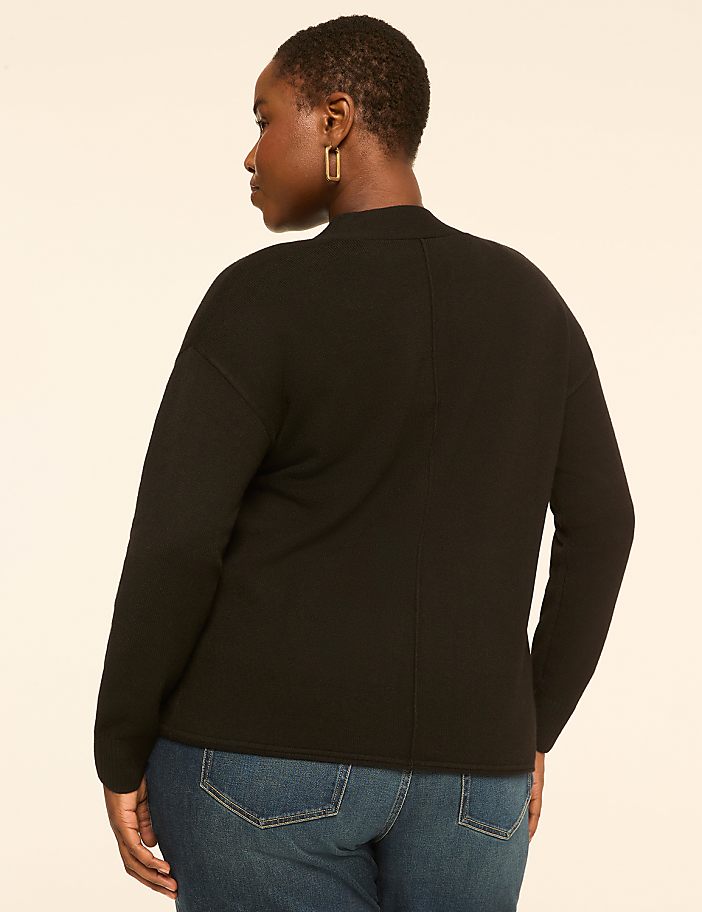 A woman in a black sweater and blue jeans stands against a plain, light-colored background.