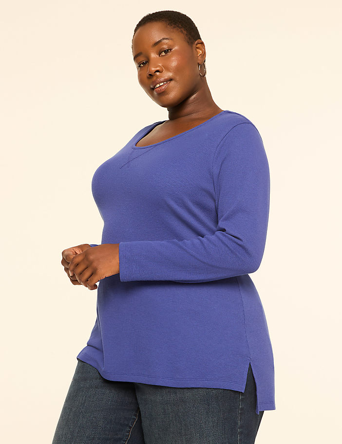 A woman in a blue long-sleeved shirt and jeans stands against a plain, light-colored background.