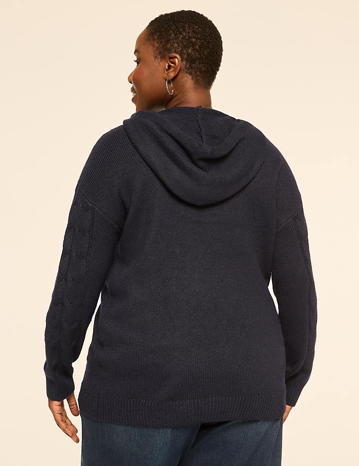 A woman in a navy blue hooded sweater and jeans stands against a plain, light-colored background.