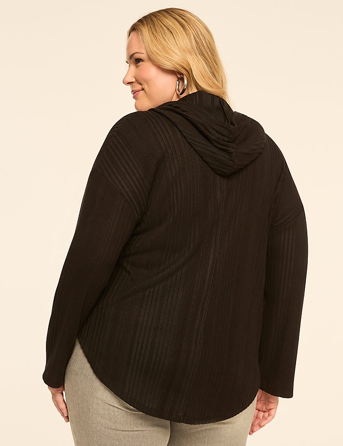 A woman in a black hooded sweater and grey pants stands against a plain beige background.