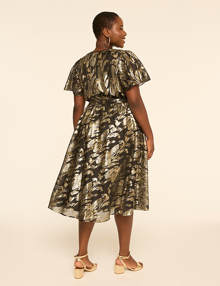 A woman in a black and gold patterned dress stands against a plain, light-colored background.