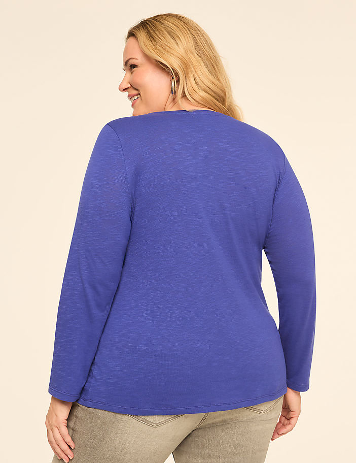 A woman in a blue long-sleeved shirt and grey pants smiles over her shoulder against a plain, light-colored background.