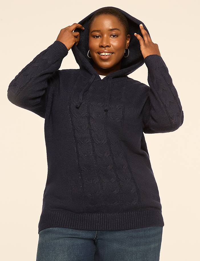 A woman in a navy cable-knit hoodie and jeans smiles as she pulls the hood up against a plain, light-colored background.