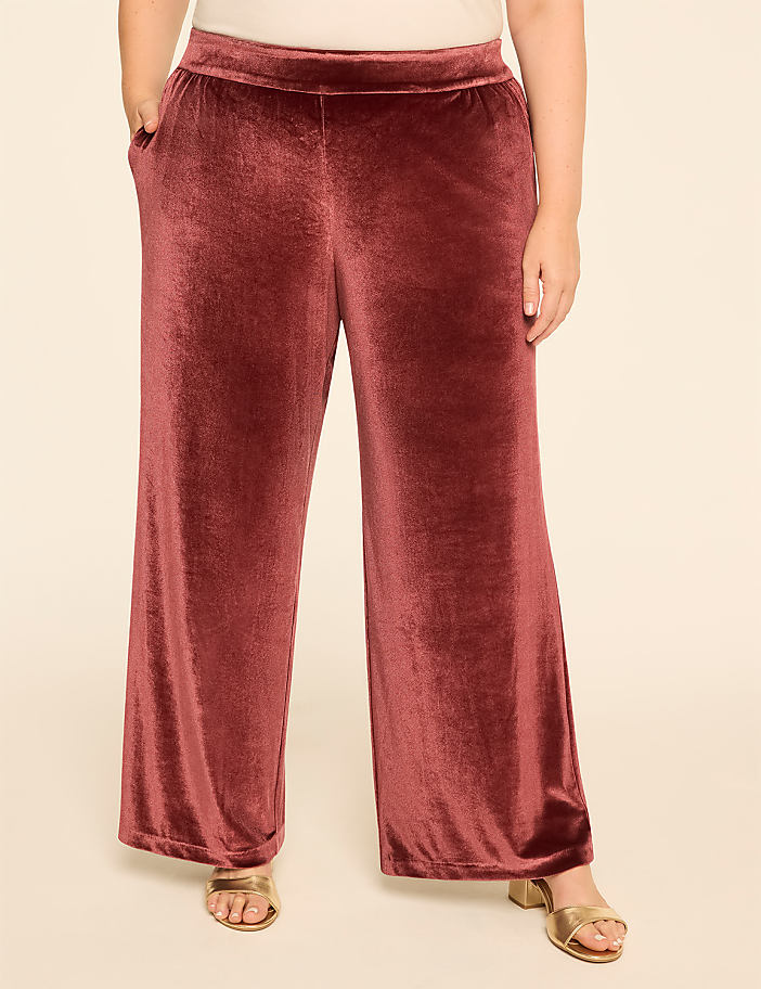 A person stands in wide-leg velvet pants with pockets and gold sandals against a plain, light-colored background.